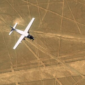 Top-down aerial view of a small white Cessna airplane flying over the straight, intersecting geometric Nazca Lines in the brown Peruvian desert.
